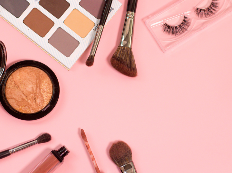 An assortment of makeup products arranged neatly over a soft pink background. The collection includes an eyeshadow palette, makeup brushes, bronzer, and lashes.