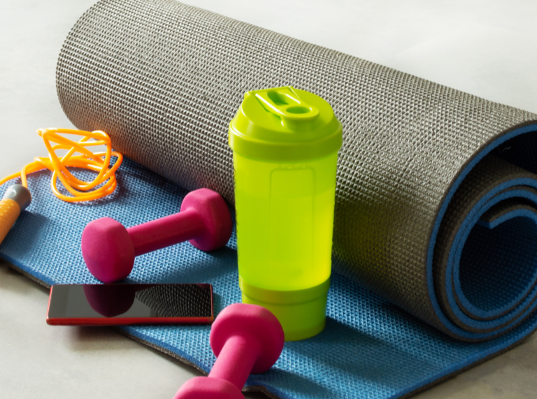 A rolled-up yoga mat is placed on the floor next to a protein shake bottle, a smartphone, and a pair of dumbbells. The items are neatly arranged, suggesting a ready-to-go workout setup.