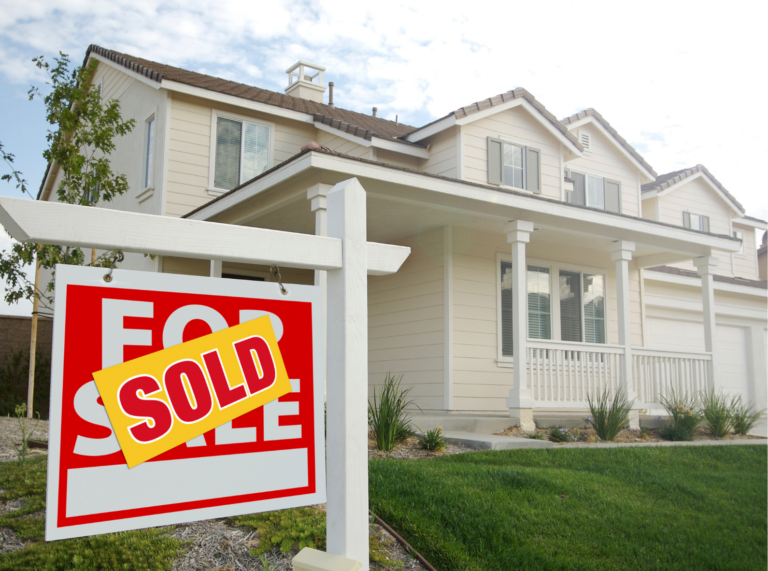 A "Sold" sign is prominently displayed in front of a beige two-story suburban house with a neatly manicured lawn. The house features a covered front porch with white railings and multiple windows, giving it a welcoming appearance. The "For Sale" sign, which now has a "Sold" label across it, is positioned on a wooden post near the front yard, indicating that the property has been purchased. The sky is partly cloudy, adding a bright and optimistic atmosphere to the scene.