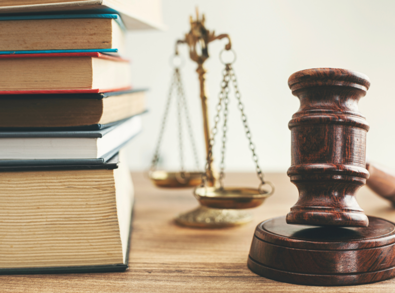 A balanced law scale centered between a stack of legal books on the left and a wooden gavel on the right, symbolizing justice, knowledge, and authority.