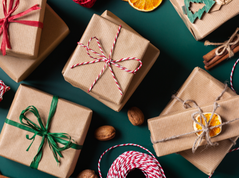 A variety of neatly wrapped gifts in brown paper with colorful twine ribbons sit against a dark green background. Some packages are tied with red and white striped twine, while others are decorated with green ribbons or natural twine. The scene includes dried orange slices, wooden ornaments, and cinnamon sticks, giving it a festive, rustic feel. This picture is to indicate presents found on writers' gift guides.