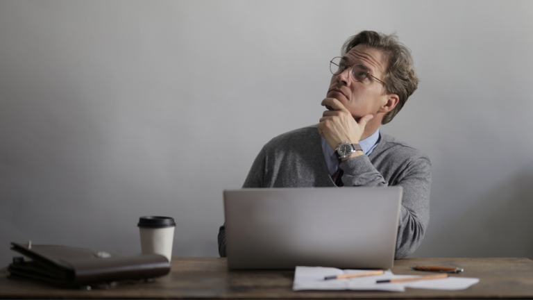 A man sits deep in thought at a table while holding a laptop. To his left sits a coffee cup. The image conveys creating thought leadership content for a public relations campaign.