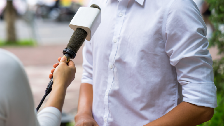 Close-up of a person in a white button-down shirt being interviewed outdoors, with a microphone held out toward them by another person.